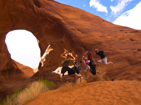 Monument Vally, United States - August 23, 2011: A group of Japanese tourists jumping for joy in the air, on the red sand in front of Monument Valley Arch. Blue sky with clouds is in background.