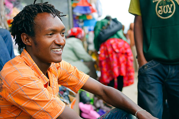 Young African man Addis Ababa, Ethiopia - July 29, 2010: A smiling young man sits on the steps of a storefront at the Merkato, the largest open air market in Africa. Another young man is walking in the background with a shirt that says JOY ethiopia city stock pictures, royalty-free photos & images