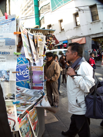 La Paz, Bolivia - May 31, 2013: Bolivian man stopping to read the latest newspapers on display outside street kiosk. People are pictured in the background