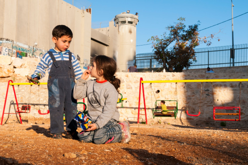 Bethlehem, West Bank, Palestinian Territories - November 2, 2010: Three Palestinian children enjoy the playground on an autumn day. The large wall and security tower in the background is part of the controversial Israeli security barrier, which in 2004 the International Court of Justice deemed illegal on account of its routing (it often veers away from the internationally recognized Green Line and deep into Palestinian territory). Three Palestinian men are also visible in the background.