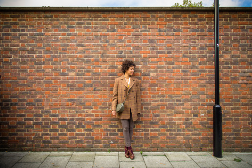 A fashionably dressed young woman stands against a brick wall, a black lamppost to her left.