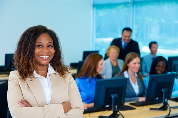 Female professor with computer lab students A smiling college instructor looking at camera with students seated behind her in a computer lab.  The computer lab has blue-themed walls and windows. professor business classroom computer stock pictures, royalty-free photos & images