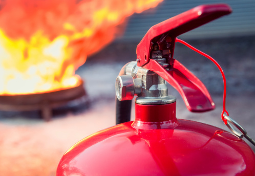 Detail of the top of a foam (AFFF - Aqueous Film-Forming Foam) fire extinguisher ready to use, with the safety pin out, and a large fire burning in the background.