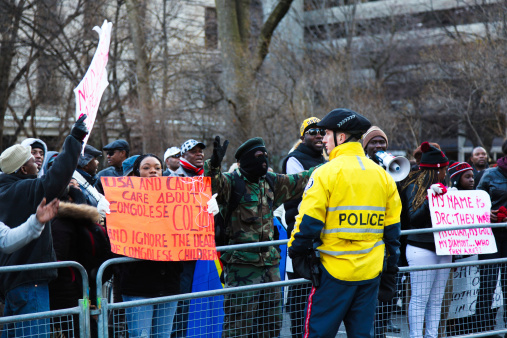 Toronto, Canada - January 07, 2012: Congolese protestors demanding safety for people in Congo behind the police barricades in downtown Toronto. A man in camouflage and a face-mask facing the police officer who is standing at the other side of the barricade.