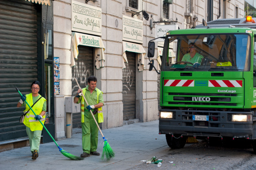 Milan, Italy - October 2, 2011: Two worker of the city owned AMSA company are sweeping a street (Corso Como, a popular spot for shopping and nightlife) with the support of a cleaning truck.