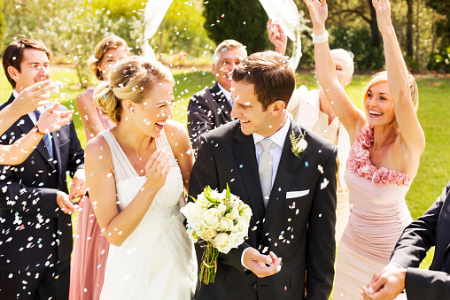 Happy guests throwing confetti on cheerful couple during wedding reception at garden. Horizontal shot.