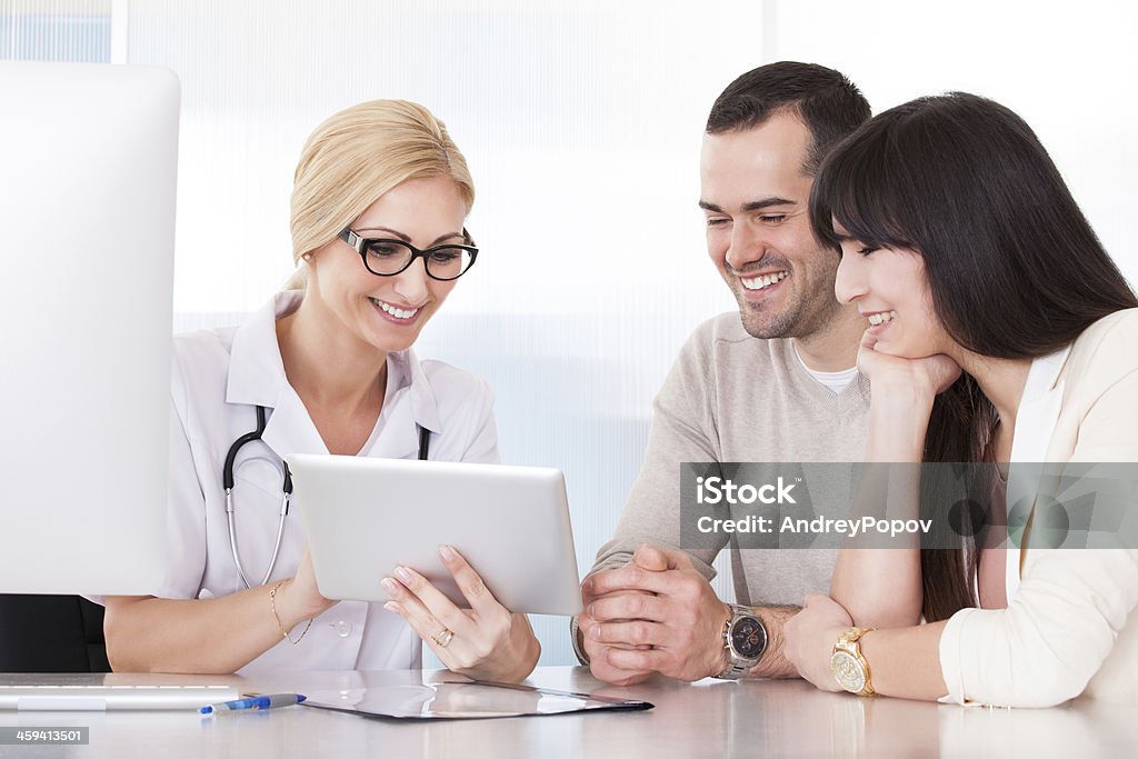 Medical professional and young couple smiling at tablet Happy Doctor Discussing With Couple In Clinic Couple - Relationship Stock Photo