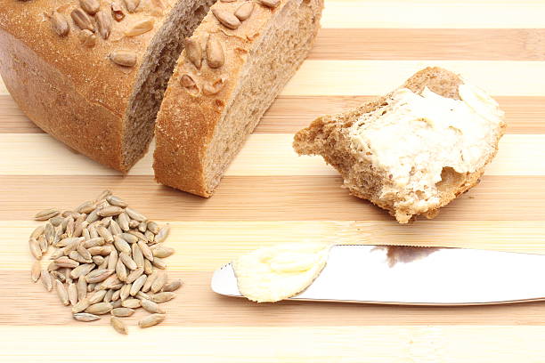 Rye grain, butter on knife and bitten slice of bread lying on wooden cutting board