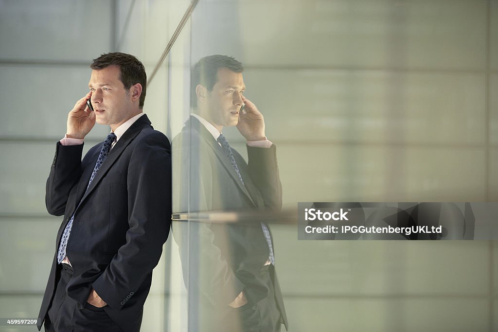 Businessman Using Mobile Phone While Leaning On Glass Wall Middle aged businessman using mobile phone while leaning on glass wall in office Adult Stock Photo