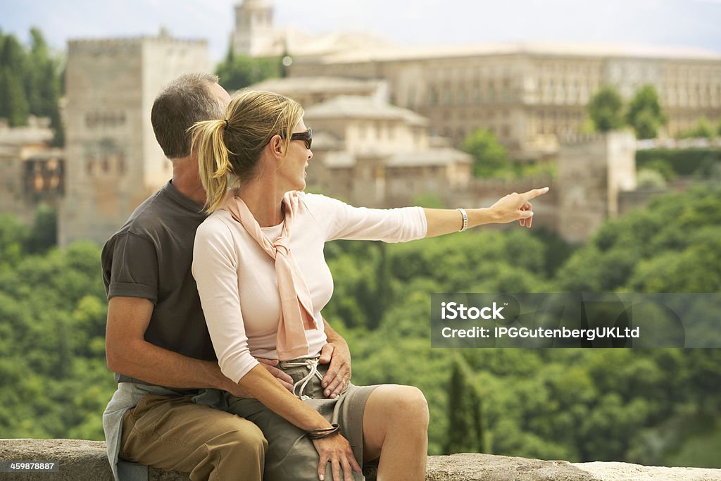 Happy Tourist Couple With Woman Pointing At View Middle aged couple sitting on wall with woman pointing at view; Granada Spain Adult Stock Photo