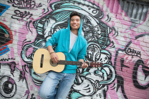 Young smiling street musician leaning on a wall with graffiti drawings and playing his guitar
