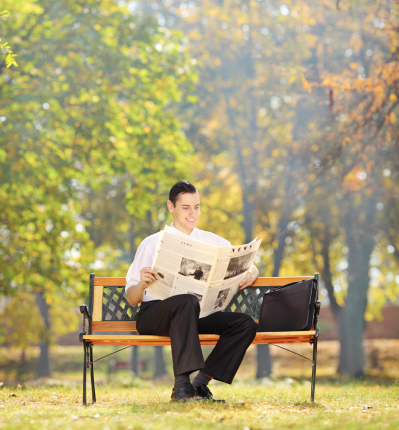 Businessman seated on a wooden bench reading a newspaper in a park, shot with a tilt and shift lens