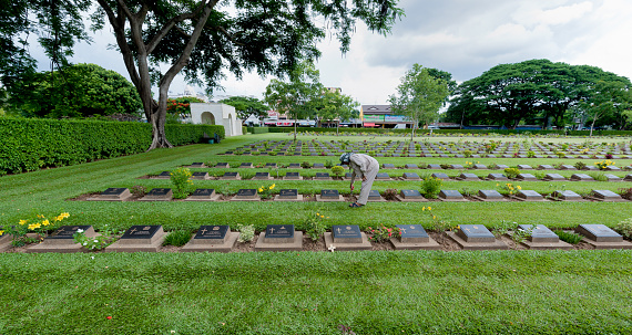 Kanchanaburi, Thailand - July 6, 2012: Thai gardener at work between the gravestones of the war cemetery of Kanchanaburi in Thailand, a memorial to the Dutch and English prisoners who died during the building of the Burma railway, the Death Railway, during the Second World War