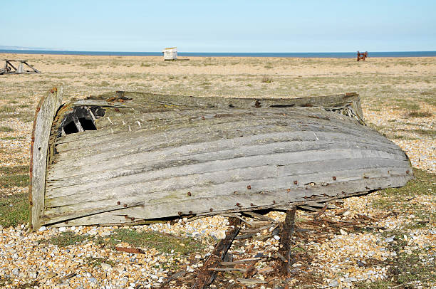 An old wooden fishing boat rotting away on the shingle beach at Dungeness, Kent.