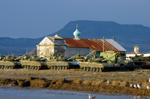 Military tanks are on the beach. Old crumbling church and cross. Blue sky and mountain background. Evening sunset