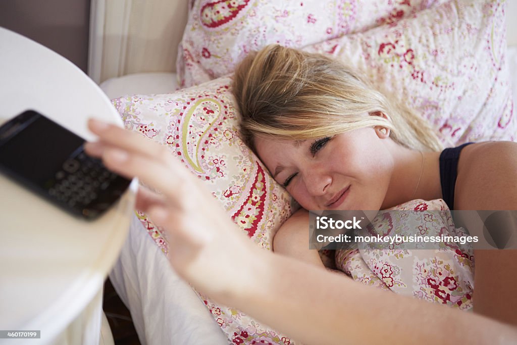 Teenage Girl Turning Off Alarm On Phone Teenage Girl Waking Up In Bed And Turning Off Alarm On Phone Bedtime Stock Photo