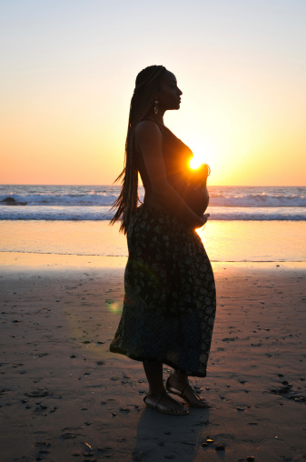 A lovely woman walking down the beach as she holds her belly. Intentionally silhouetted exposing for the background and with a little bit of lens flare. Vertical image with copy space.