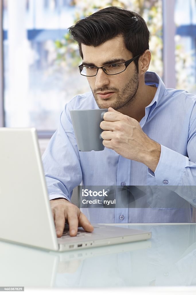 Businessman working at desk Businessman sitting at desk, working on laptop computer, drinking coffee.. 20-29 Years Stock Photo