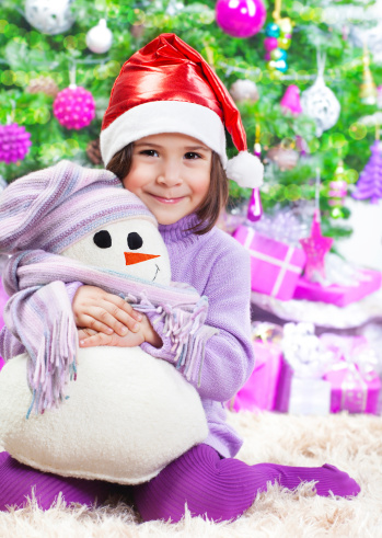 Cute little girl sitting on the floor under big decorated Christmas tree, wearing red Santa hat, holding in hands white snowman soft toy, New Year holidays