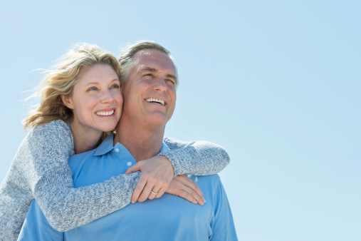 Low angle view of happy mature woman embracing man from behind while looking away against clear sky