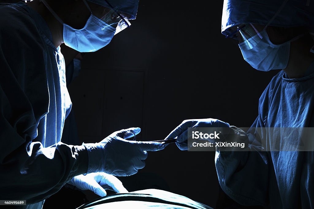 Surgeons passing surgical equipment in the operating room, dark Two surgeons working and passing surgical equipment in the operating room, dark Operating Room Stock Photo