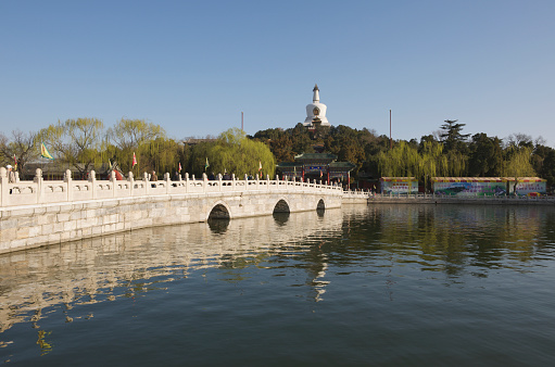 Beijing, China - April 4, 2012: It is early morning in the Beihai Park - and clear blue sky - in the spring. We see a white marble bridge leading from the south entrance to the Qionghua Island, a large multi-colored wooden paifang (also called pailou - a traditional Chinese archway) and on top of the hill, the Bai Ta (White Pagoda) a 40 m high stupa. People are practicing Tai Chi under the weeping willow trees.