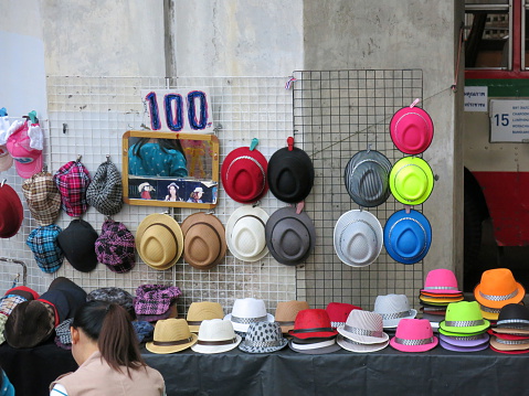 Bangkok, Thailand – December 27, 2013:  A view of street vendor selling hats on the sidewalk in Bangkok. Street vendors sell goods on a sidewalk in Bangkok, Thailand. A blind man sings to beg for money.  Pedestrians walk on the sidewalk.