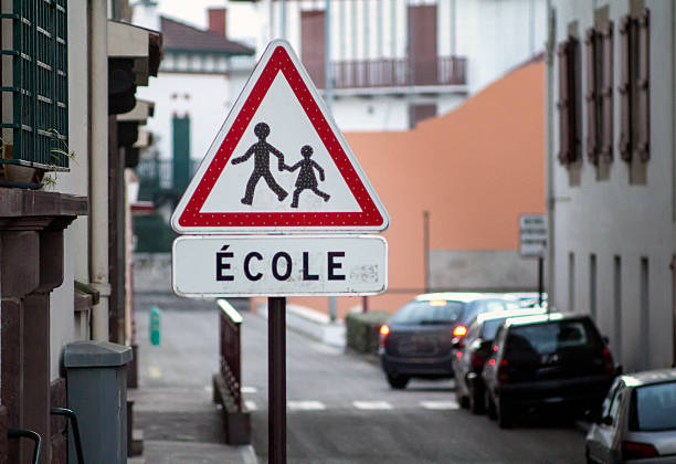 French school crossing sign A French sign warning of students crossing the road. Show with a shallow depth of field. License plate numbers of the out of focus cars in the background have been edited out. Typical basque architecture is visible in the background. Shot in St Jean de Luz in the Pays Basque region of France. hazard sign photos stock pictures, royalty-free photos & images