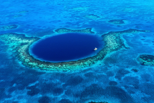 An aerial view of a boat moored in the Great Blue Hole off the coast of Belize. The azure seas of the Caribbean envelope the deep blue of the Great Blue Hole. A coral reef surrounds the geologic wonder.