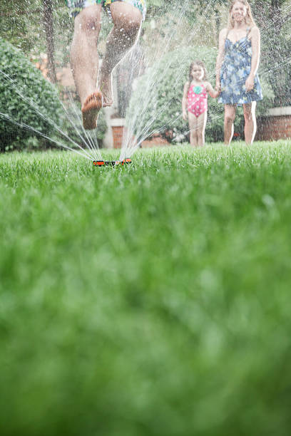 istock Father jumping through a sprinkler, mother and daughter watching 460773193