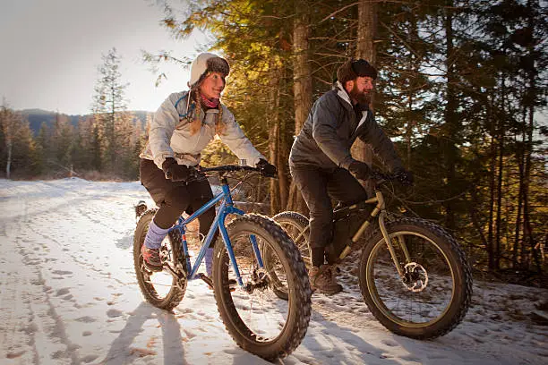 A man and a woman are riding fatbikes on a snow covered trail in northern Idaho. The sun is low in the sky and giving a warm glow to the scene.