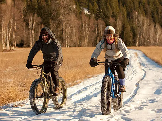 A man and a woman are riding their snowbikes along a snow covered trail on a sunny day. Shot near some woods in northern Idaho on a crisp winter day.