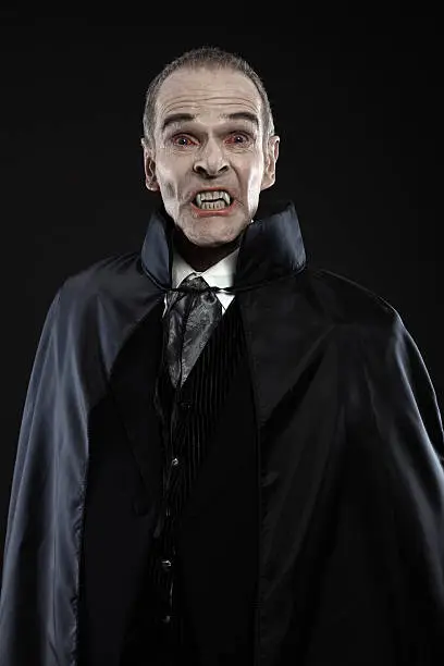 Dracula with black cape showing his scary teeth. Vamp fangs. Studio shot against black.