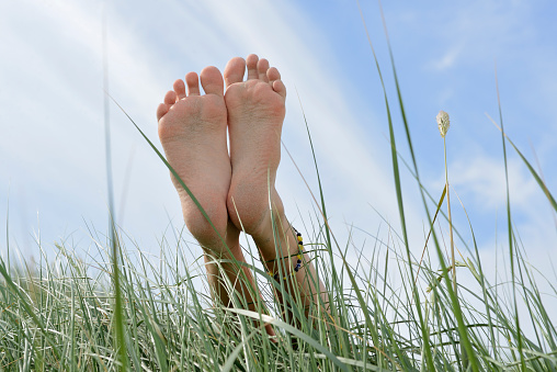 Barefoot on the sand dune. Slim feet and legs of a caucasian girl. Feet are stretched over american beachgrass or marram grass. Light blue sky in the background. Taken from low angle view in summer on a sand dune in Baltrum Island, Ostfriesland, German North Sea Region, Lower Saxony, Germany, Europe.