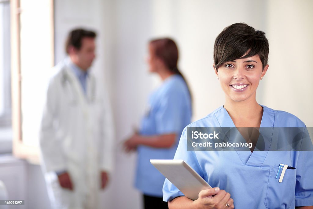 Lovely latin nurse on blue uniform standing Portrait of a lovely latin nurse on blue uniform standing and smiling on medical team background Adult Stock Photo