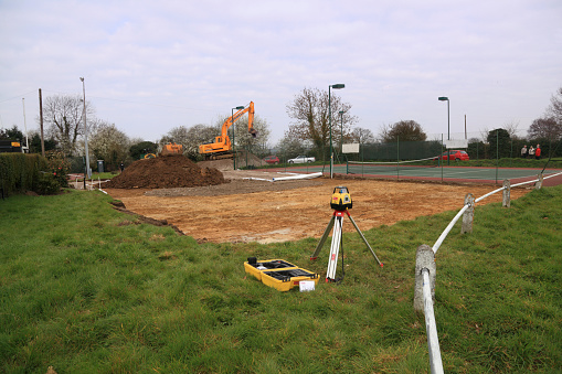 Epping, United Kingdom - March 22, 2011: The first stage of building a new tennis court is seen. The topsoil has been excavated and is piled up at one end of the new court. An excavator is parked on top of the new hardcore foundation material, waiting for a truck to remove the spoil A theodolite in the foreground has been used to establish building levels.