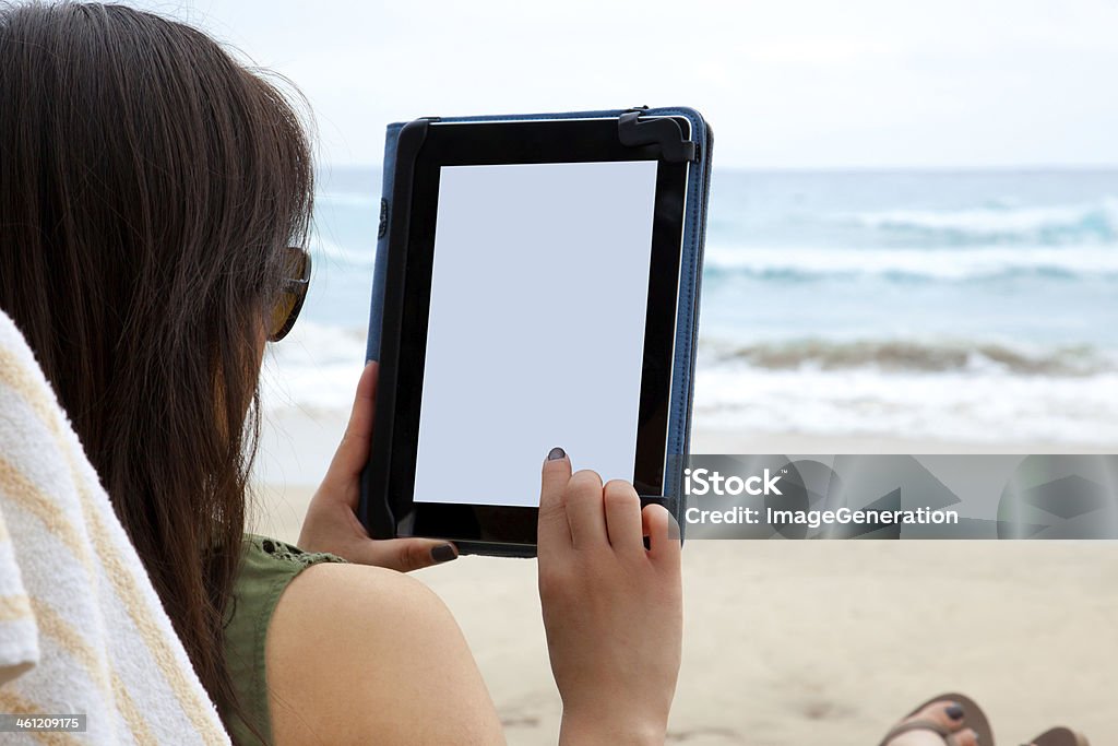 Woman sitting on the beach enjoying the view of her tablet A woman uses a tablet device while on the beach, symbolizing the ability to blend work and rest while working from anywhere. Beach Stock Photo