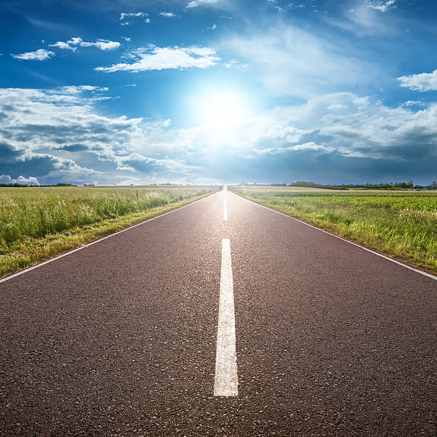 Asphalt road leading to horizon with grass and blue sky Driving on an empty road towards the sun empty road with trees stock pictures, royalty-free photos & images