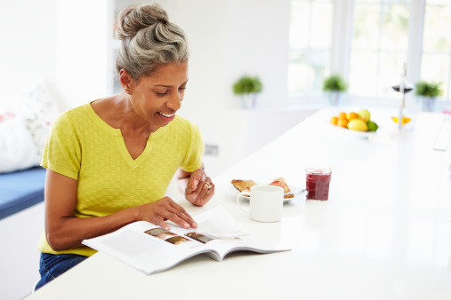 Woman Eating Breakfast And Reading Magazine In Kitchen