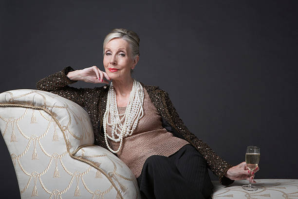 Elegant Senior Woman On Chaise Lounge With Champagne Portrait of an elegant senior woman sitting on chaise lounge with champagne against black background chaise longue stock pictures, royalty-free photos & images