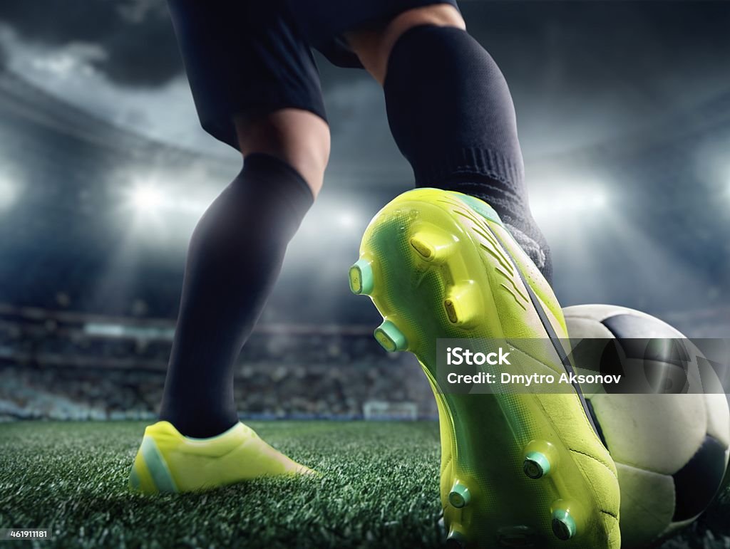 Close up of a soccer player's foot in a stadium A ground shot of a soccer player's foot wearing a lime-green shoe kicking a soccer ball.  The player is wearing black socks and black shorts.  In the background, out of focus, is a stadium with an audience.  Bright white lights illuminate the scene under a stormy sky. Close-up Stock Photo