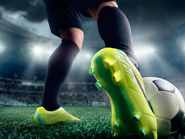 Close up of a soccer player's foot in a stadium A ground shot of a soccer player's foot wearing a lime-green shoe kicking a soccer ball.  The player is wearing black socks and black shorts.  In the background, out of focus, is a stadium with an audience.  Bright white lights illuminate the scene under a stormy sky. cleats stock pictures, royalty-free photos & images