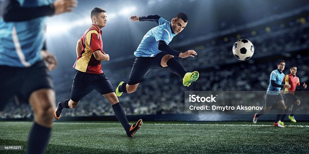 Soccer players in stadium Mid-air action shot of professional footballers tackling on pitch during game in full floodlit stadium Soccer Competition Stock Photo