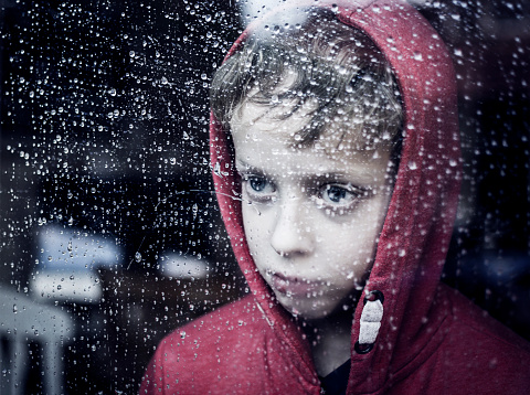 Little child looking through window. Raining outside. Depressed or sad. Can't play or bullied boy.