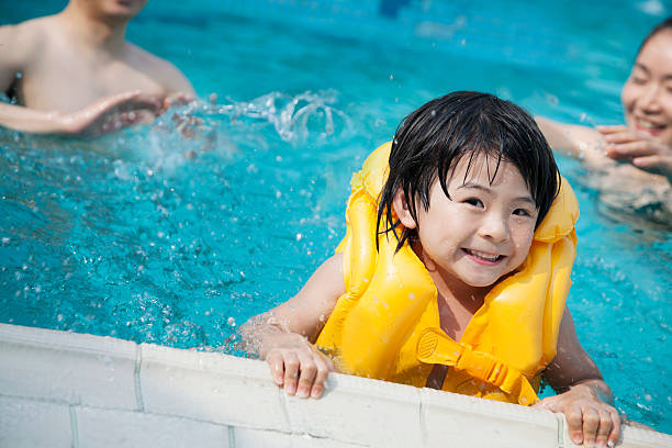 Young boy wearing flotation device holds onto edge of pool Portrait of smiling son in the water and holding onto the pools edge with family in the background life jacket stock pictures, royalty-free photos & images