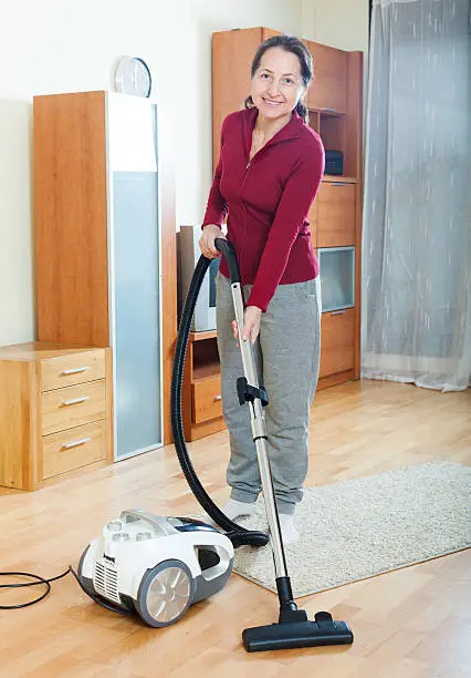 Happy mature woman vacuuming with vacuum cleaner on parquet floor