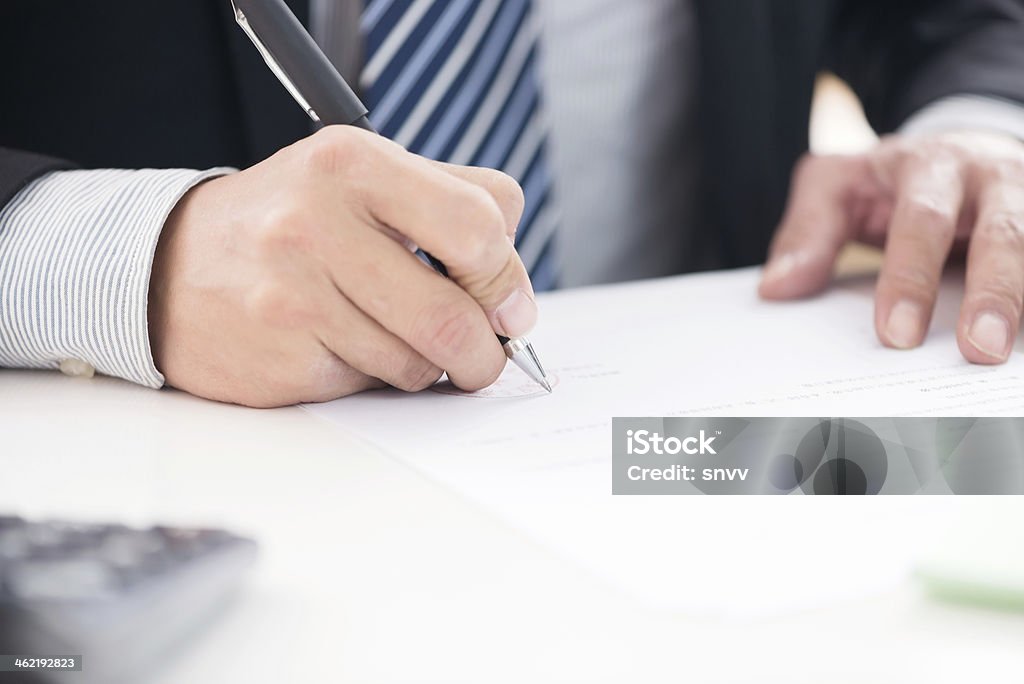 Close up of businessman signing a contract Businessman sitting at office desk signing a contract with shallow focus on signature. Adult Stock Photo