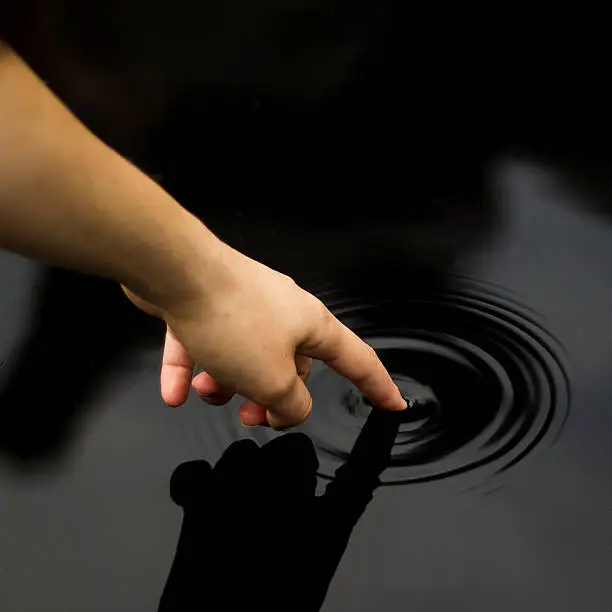 High Angle Shot of a Finger Touching Black Water Causing a Ripple with a Shadow of the Hand Reflected on the Water.