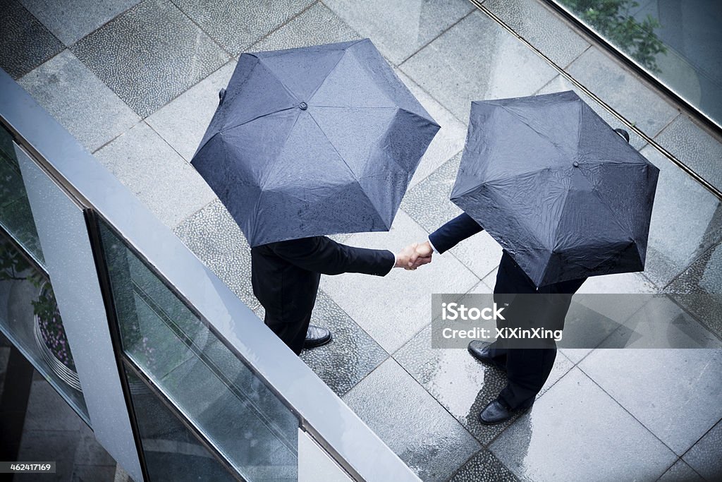 Dos hombres de negocios estrechándose las manos sosteniendo sombrillas y en la lluvia - Foto de stock de Paraguas libre de derechos