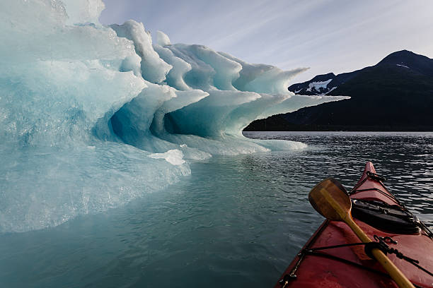 Paddler pauses to look at Melting Iceberg Paddler lashes paddle to kayak to pause journey to enjoy the splendor of the shapes of the  melting Iceberg in summertime Alaska's Kenai Fiords National Park seward alaska stock pictures, royalty-free photos & images